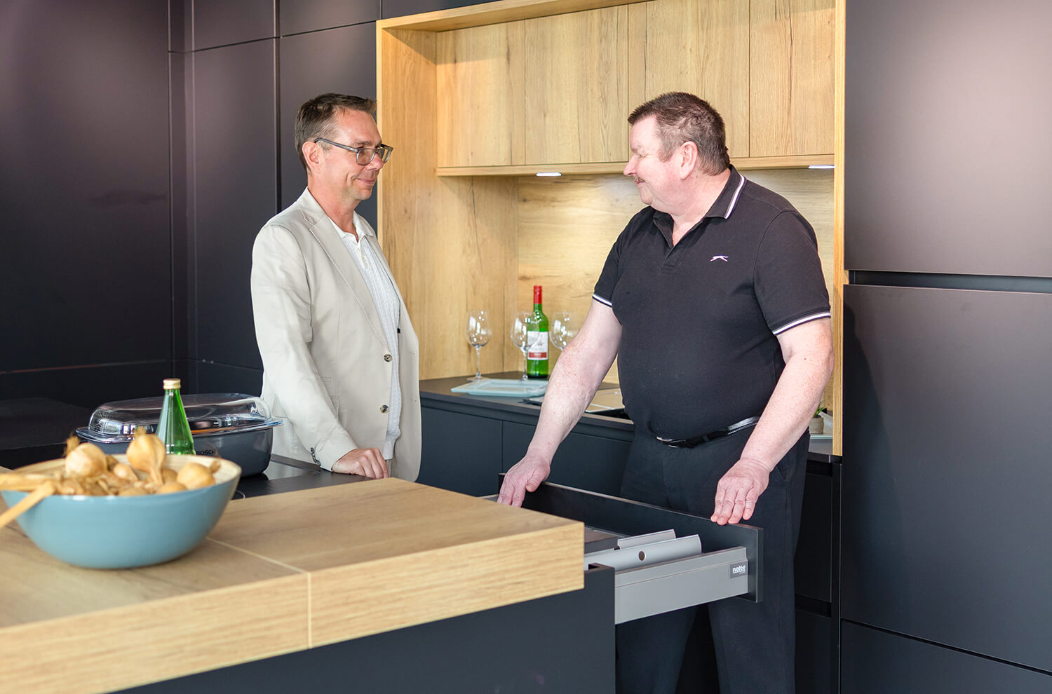 Zwei Männer unterhalten sich in einer modernen Küche mit schwarzen Fronten und Holzakzenten über Küchendetails.