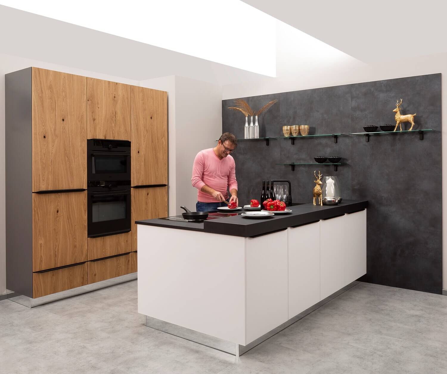 Helle Designküche von Bauformat mit Holzfronten und schwarzer Arbeitsplatte, Mann beim Kochen vor dunkler Rückwand.